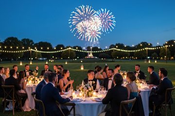 Réception de mariage en plein air au crépuscule, avec des invités élégamment vêtus observant un feu d’artifice lointain dans un grand parc.